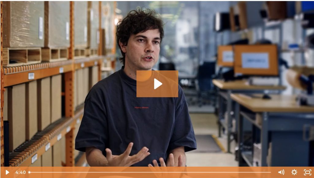 A person speaks while seated in an industrial workspace, surrounded by shelves and desks with computer monitors displaying a WMS inventory management system. Video player controls are visible at the bottom.